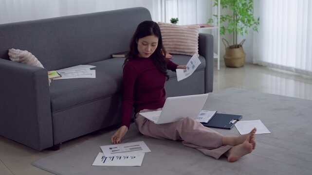 Young Asian woman reviewing financial documents and laptop while working from home sitting on living room floor
