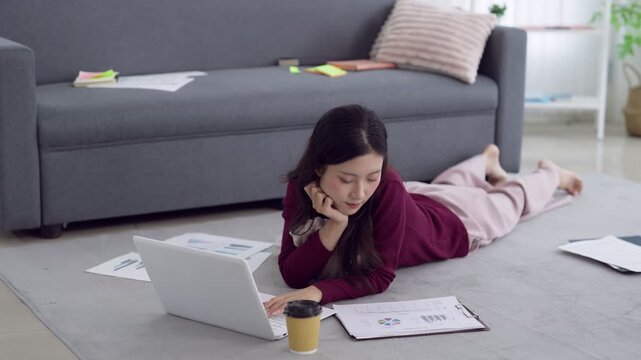 Young Asian gen z woman working remotely on laptop lying on living room floor with business reports and coffee