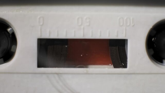 Macro close-up of a white audio cassette playing in a deck with a slow zoom out. Black spools rotate to wind the magnetic tape behind the vintage counter window.
