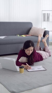 Young Asian woman working remotely on laptop and business reports lying on floor at modern home