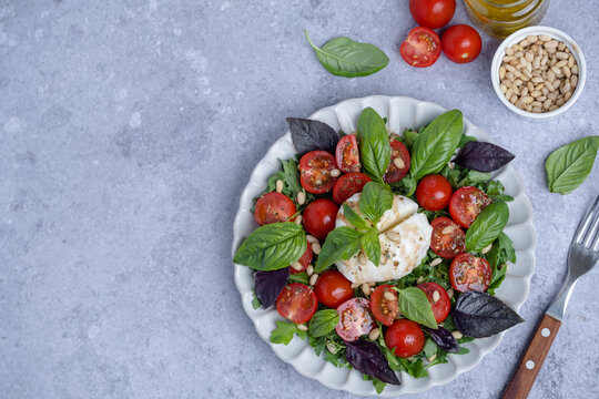 Fresh burrata salad served with ripe cherry tomatoes, mixed greens, fresh basil leaves, and toasted pine nuts on a gray background. Healthy eating concepts, and Italian cuisine. top view