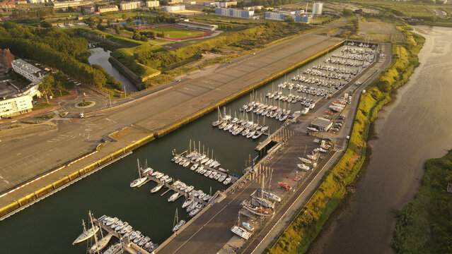 Aerial view of the marina in Calais with numerous sailboats docked in rows, adjacent to a road, green fields, and a sports stadium in the background, Calais, Hauts-de-France, France.