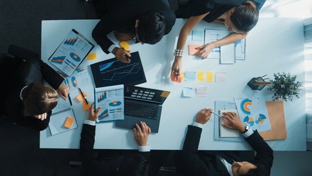Top down aerial view of diverse business people discuss and brainstorm marketing idea while looking at financial graph at table with marketing document and sticky notes scatter around. Directorate.