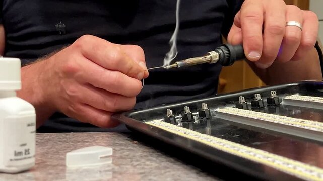 Hands work diligently on a circuit board, soldering tiny components with precision. Smoke rises as the soldering iron connects parts, showcasing craftsmanship.