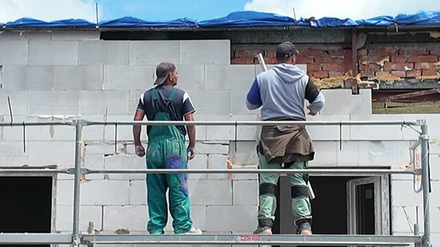 Construction worker on scaffolding building a wall with concrete blocks. A builder in protective gear works on a new structure, meticulously placing concrete blocks and preparing for window installati