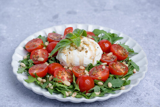 A round plate holds a colorful salad made with cherry tomatoes, herbs, and soft cheese. Pine nuts are sprinkled on top, creating a fresh and tasty dish in a kitchen.