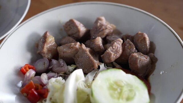 Close-up of tender beef chunks served with shallots, chili, cabbage, cucumber, and sauce in a bowl, traditional Asian food footage with natural light.