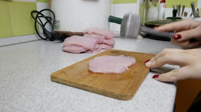 Female hands use a metal meat tenderizer to pound a raw pork loin on a wooden cutting board. The close-up shot captures the domestic food preparation process in a home kitchen.