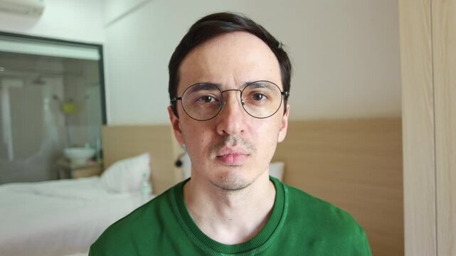Caucasian man with eyeglasses looking directly at the camera, transitioning through a range of expressions from neutral to a subtle smile while staying in a modern interior bedroom setting