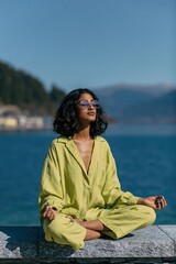 Fototapeta premium Woman in lime green relaxed suit meditating by a lake with sunglasses for yoga background and wellness poster