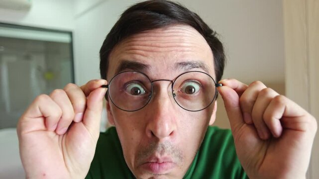 Funny and slightly crazy man squinting, putting on glasses, and making a comical wide-eyed WOW expression of surprise. Eccentric guy amazed by clear vision and perfect eyesight in front of the webcam