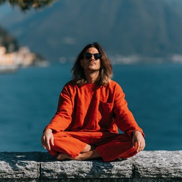 Young man practicing meditation by the ocean in bright orange outfit for wellness poster and calming background use