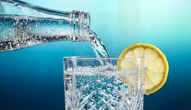 Sparkling water poured from glass bottle into crystal tumbler with lemon slice, refreshing beverage with bubbles, healthy drink, studio shot.