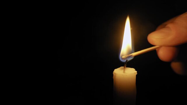 Hand holds a burning match to light a single wax candle wick. Bright flame illuminates the dark, creating a warm, intimate glow on a pure black background.