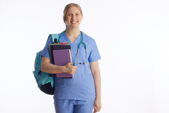 A young woman in medical scrubs carries a stack of notebooks and a pen, representing the academic side of nursing school, exam preparation, and clinical studies.
