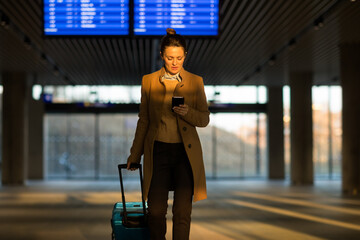 Fototapeta premium A professional woman in a tan coat checks her smartphone while standing with a blue rolling suitcase in a modern railway station terminal.