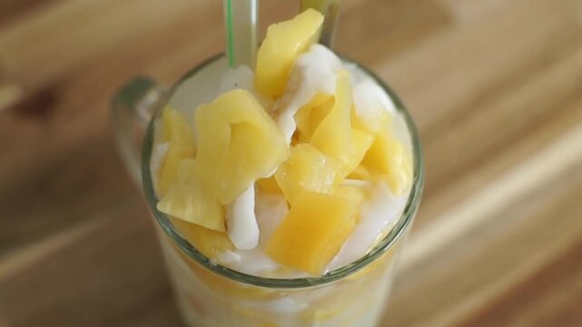 Close-up of tropical pineapple dessert in a glass with creamy topping, fresh fruit chunks, and soft natural light on a wooden table.