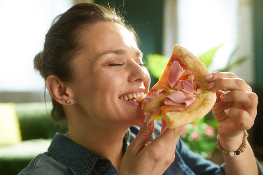 A woman in a denim shirt smiles with her eyes closed while enjoying a slice of ham pizza, capturing a relaxed and satisfying lunch break at home.