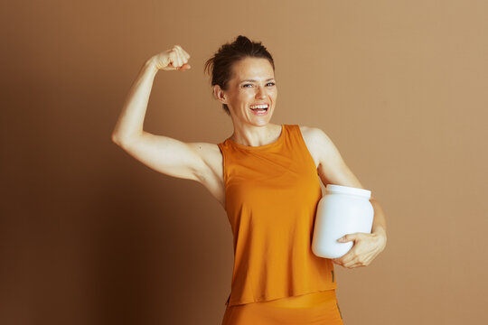 A strong woman in her 30s, hair in a bun, flexes her bicep while holding a large white container and smiling broadly. She embodies fitness, nutrition, and athleticism against a brown background.