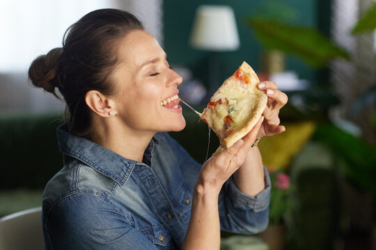 A relaxed woman in a denim shirt prepares to take a bite of a cheesy pizza slice in a cozy indoor setting, highlighting casual home dining and comfort food choices.