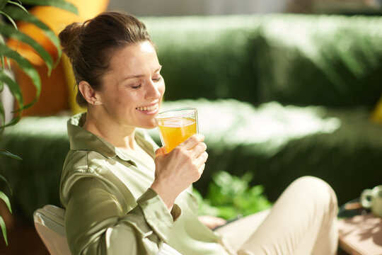 A woman enjoys a peaceful moment at home with her eyes closed and a gentle smile, holding a glass of orange juice in a sunlit room for a mindful wellness routine.
