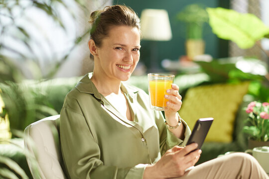 A cheerful woman in her 30s sits in a bright room, holding a glass of orange juice and a smartphone while looking warmly at the camera during her morning routine.