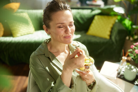 A woman finds peace while eating a fresh fruit salad with her eyes closed, enjoying a quiet moment of self-care and healthy nutrition in her living room.