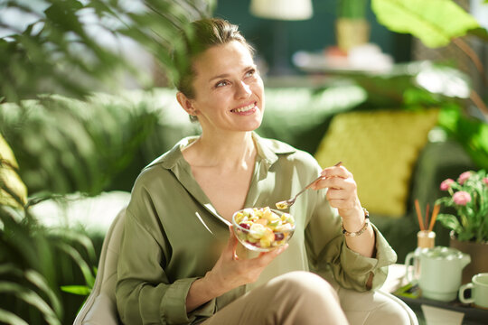 Looking contentedly upward, a woman sits in her living room with a bowl of fresh kiwi and melon salad, surrounded by green plants and soft natural light.