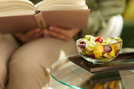 A glass bowl of colorful fruit salad rests on a dark wooden tray next to an open book, symbolizing a peaceful home snack break and a healthy self-care routine.