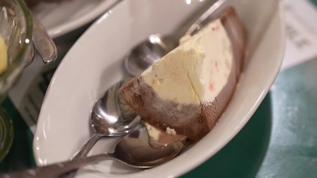 Close-up of layered ice cream dessert in a white bowl with chocolate and vanilla flavors, served with spoons, creamy frozen treat footage with soft restaurant lighting.