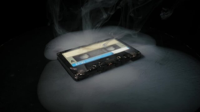 Old audio cassette tape with an EF90 label sits on a black surface as dense fog clears. Dramatic spotlight lighting illuminates the retro object against a pure black background.