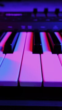 Finger presses keys on an electronic synthesizer keyboard. Close-up shot is illuminated by vibrant pink, purple, and blue neon lights, creating a moody, retro atmosphere.