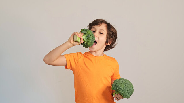 Eye level portrait of a playful boy in an orange shirt pretending to bite fresh broccoli against a plain wall. Child nutrition, picky eating, and healthy snacks shape everyday family food choices.
