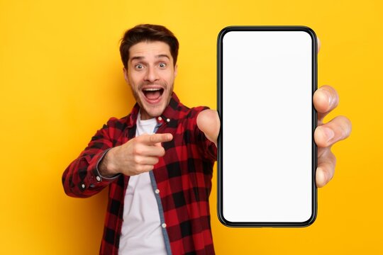 Smiling Young Caucasian Man Pointing At Smartphone With Blank Screen, Expressing Excitement In Studio Setting With Yellow Background, Perfect For Mockup And Advertising Concept.
