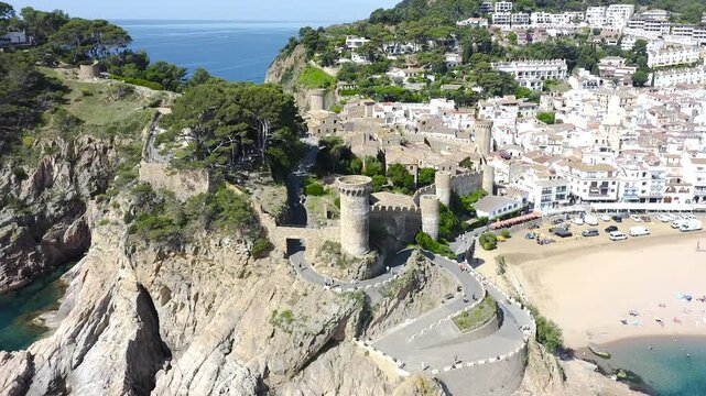 Aerial view of Tossa de Mar, a historic Costa Brava town crowned by its medieval walls, overlooking a beautiful sandy beach and turquoise Mediterranean waters. 
