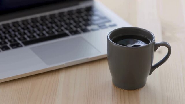 Minimalist black coffee in grey ceramic mug on wooden desk, laptop computer in blurred background, modern remote work office setup