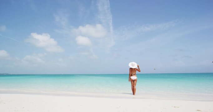 Beach bikini woman standing by ocean enjoying paradise beach resort. Young lady wearing white bikini and and elegant sun hat during summer vacation. Idyllic beach view sunny day.