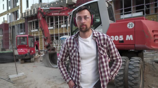 Portrait of proud builder at construction site. Constructor with excavator prepared to start building new house. Handsome construction worker standing in an outdoor area with heavy machinery. Engineer