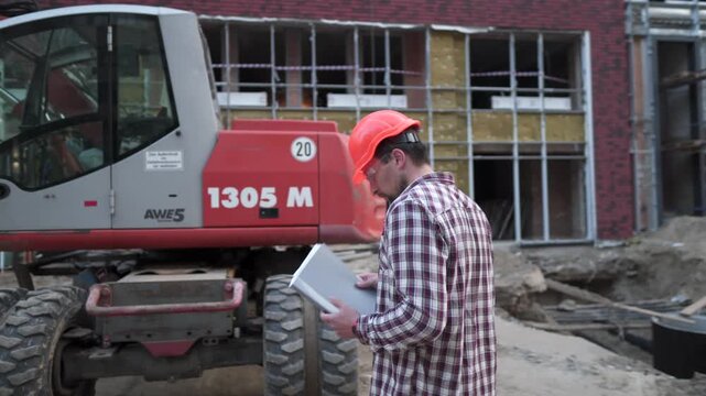 Young construction manager checks the technical documentation while on construction site near heavy construction equipment tractor or excavator. Foreman checking report.