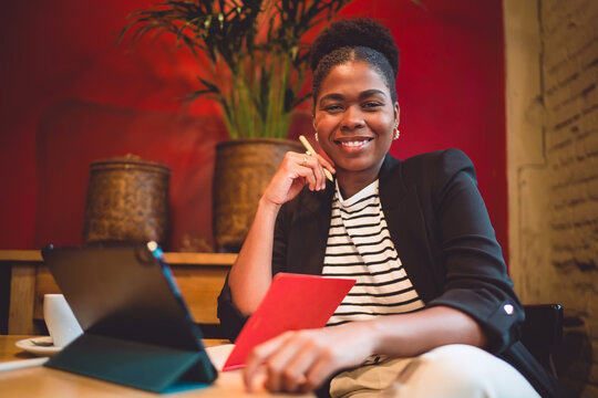 Smiling Black woman holds stylus and red notebook at cozy caf&eacute; table with tablet, representing confident freelance work, digital creativity, and joyful focus in modern business setting.