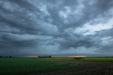 Fototapeta premium Dramatic dark storm clouds sweep over a flat rural farmland as a thin band of sunset light glows on the distant horizon