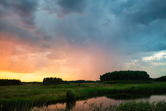 Lightning storm over river meadow with thunderclouds and reflective water in evening
