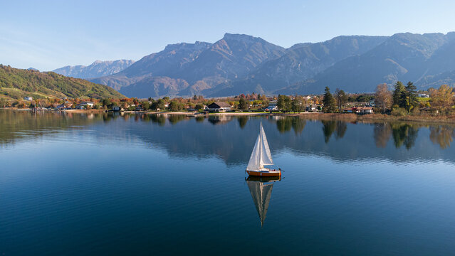 Aerial 4K drone view of a sailboat on calm Lake Caldonazzo, Italy. Features perfect mirror reflections of the scenic Trentino mountains and autumnal shoreline on still blue water.