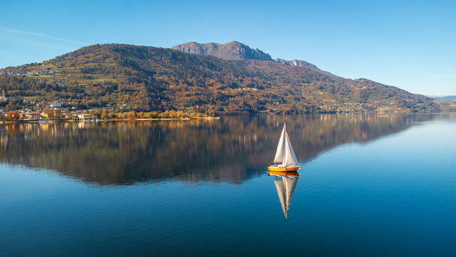 Aerial 4K drone view of a sailboat on calm Lake Caldonazzo, Italy. Features perfect mirror reflections of the scenic Trentino mountains and autumnal shoreline on still blue water.
