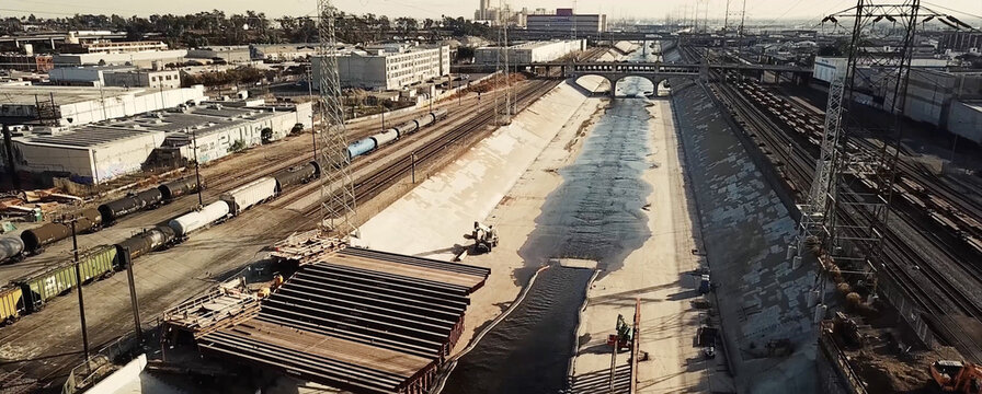 Aerial view of the concrete-lined Los Angeles River channel with freight trains on adjacent tracks, industrial buildings, and a bridge in Los Angeles, California, United States.