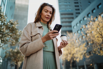 Fototapeta premium Close up of mature Caucasian female looking at smartphone screen holding disposable cup. Modern woman checking mobile app notifications or reading news in city sunlight. Authentic representation.