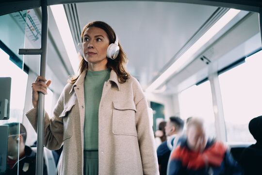 Middle aged Caucasian woman standing in moving tram, wearing wireless headphones, looking out window thoughtfully, public transport commuting, urban lifestyle, authentic passenger portrait.