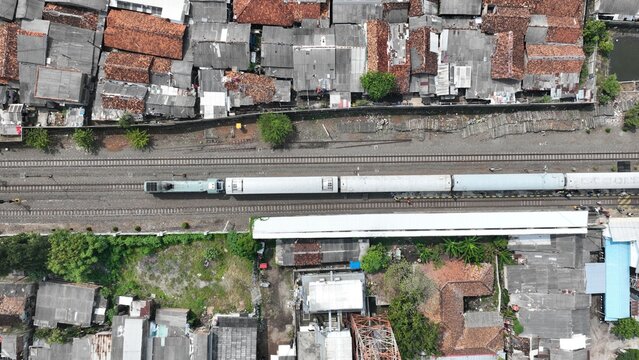 Aerial view of a passenger train traveling along tracks through the dense urban landscape of Serang with terracotta rooftops and lush greenery in Kecamatan Serang, Banten, Indonesia.