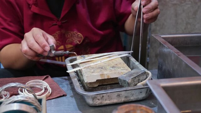 silversmith heats silver strip with blowtorch on stone block. skilled silversmith uses tweezers to handle hot metal. traditional silversmith cools heated silver in water bath to harden metal piece.