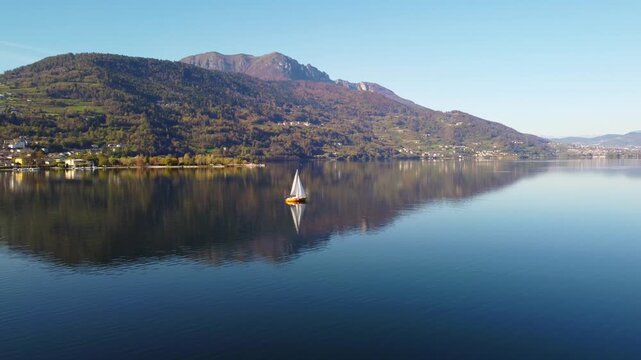 Aerial 4K drone view of a sailboat on calm Lake Caldonazzo, Italy. Features perfect mirror reflections of the scenic Trentino mountains and autumnal shoreline on still blue water.